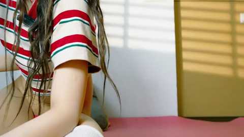 Media: Video of a person with long, wavy brown hair, wearing a red and white striped shirt, kneeling on a pink bed, in a room with white walls and a wooden door.