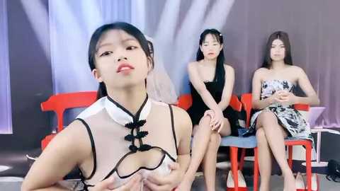 Media: Video of three Asian women with long black hair in traditional Chinese dresses on red chairs. Background is a grey curtain with white lights.