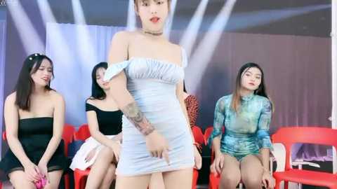 Media: Video of four Asian women, including a transgender woman with long hair in a tight, strapless light blue dress, standing in a modern, minimalist room with red chairs.