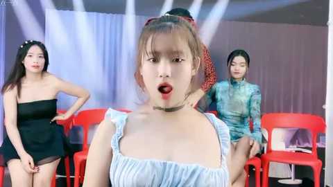 Media: Video of three young Asian women in a studio setting. Front woman has light skin, red lipstick, and wears a white blouse. Background includes a blue curtain, red chairs, and two other women in black and teal outfits.