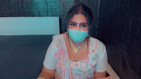 Media: Video of a woman with medium brown skin, wearing a pink floral saree, blue mask, gold necklace, sitting on a gray carpet in a dimly lit room with black curtains.