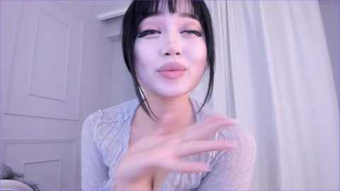 Media: Video of an East Asian woman with light skin and straight black hair, wearing a sheer, light blue lace top, applying white makeup to her face. Background includes white curtains and a door.