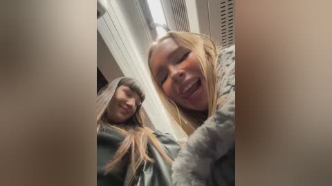 Media: Video of two young women with long hair and light skin, smiling and hugging closely. They wear winter coats and scarves, standing in a brightly lit indoor hallway.