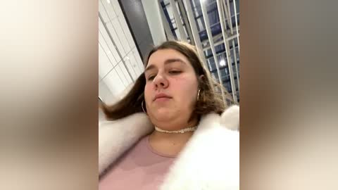 Media: A video of a young woman with light brown hair and a soft, pink sweater, standing in a tiled room with vertical blinds, appearing relaxed with eyes closed.