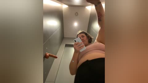 Media: A video of a plus-sized woman with short dark hair, wearing a pink sports bra and black pants, taking a selfie in a dimly lit, narrow hallway with gray walls and a red dildo mounted on a wall.