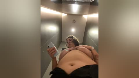 Media: Video of a woman with light skin, medium build, and long brown hair, partially lifting her shirt to reveal her midriff. She holds a smartphone, capturing a selfie in a narrow, dimly lit hallway with beige walls and fluorescent lighting.