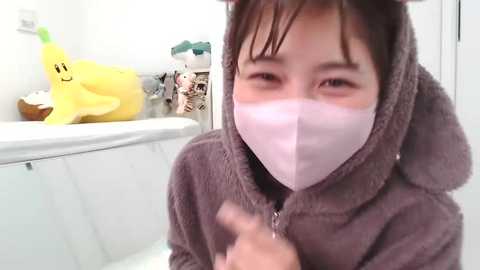 Media: Video of an Asian child in a brown, fuzzy, bear costume with a white mask, giving a thumbs-up in a bathroom with a yellow banana plush toy and toiletries.