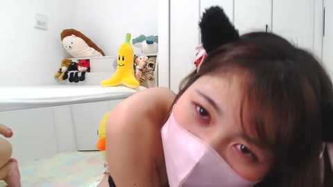 Media: Video of an Asian woman with long black hair and a pink mask, wearing a black dress, lying on a pastel-colored floor. Background includes white walls, a toy basket with plush toys, and a closet.