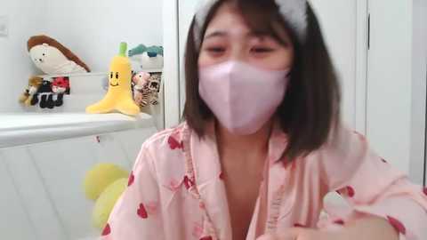 Media: Video of an Asian woman with straight black hair, wearing a pink mask, pajama top, and a white headband, sitting in a white room with stuffed toys.