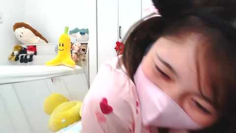 Media: Video of a young Asian girl with dark hair wearing a pink mask, surrounded by stuffed toys, including a yellow banana, in a white room.