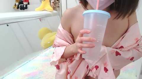 Media: A video of a woman in a pink satin robe with heart patterns, holding a clear plastic cup with a pink straw, in a bathroom with white walls, yellow stuffed toys, and a yellow bath mat.