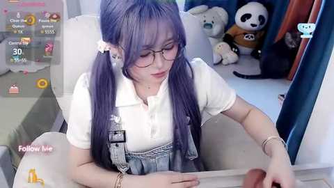 Media: Video of an Asian woman with long purple hair in pigtails, wearing glasses, a white polo shirt, and denim overalls, playing a video game. Background includes a plush panda and stuffed toys.