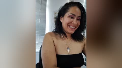 Media: Video of a smiling woman with shoulder-length black hair, wearing a strapless black top, sitting indoors with white blinds in the background.