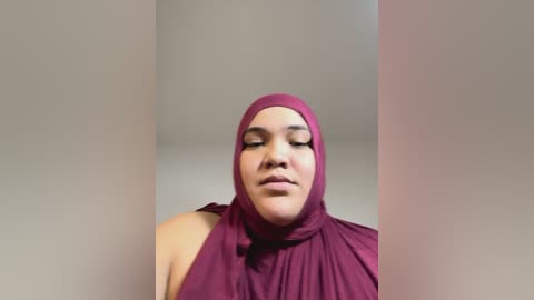 Media: Video of a woman with light brown skin, wearing a maroon hijab and dress, eyes closed in a serene pose, against a plain beige background.