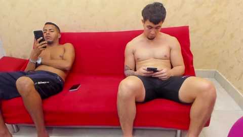 Video of two shirtless men, one wearing black shorts, the other in navy shorts, sitting on a red couch, absorbed in their smartphones, against beige wallpaper.