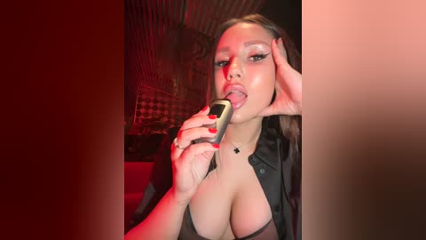 Media: Video of a young woman with fair skin, large breasts, and dark hair, wearing a black shirt, licking a sex toy with red lighting.