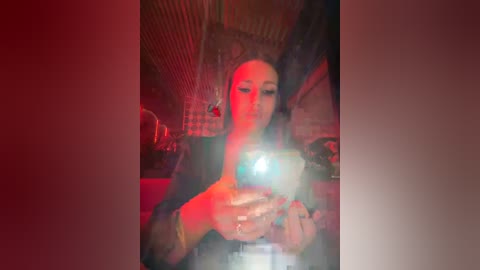 Media: Video of a woman with long, dark hair, wearing a black top, taking a selfie with a phone in a dimly lit room with red and blue lighting.