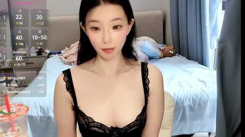 Media: Video of a young Asian woman with long black hair, wearing a black lace bra, sitting on a bed with light blue sheets in a modern bedroom.