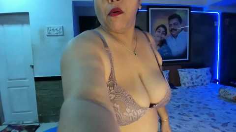 Media: Video of a middle-aged woman with fair skin and red lipstick, wearing a beige lace bra, standing in a dimly-lit bedroom with a large family portrait on the wall.