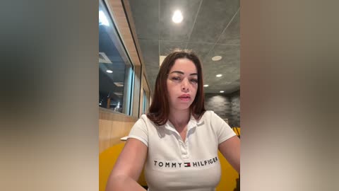 Media: A video of a young woman with medium skin tone and brown hair, wearing a white Tommy Hilfiger polo shirt, sitting indoors with a modern, industrial-style ceiling and yellow chairs.