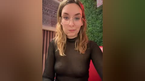 Media: Video of a Caucasian woman with light skin and shoulder-length wavy blonde hair, wearing glasses and a sheer black top that reveals her small breasts. She stands indoors with greenery and red decor in the background.