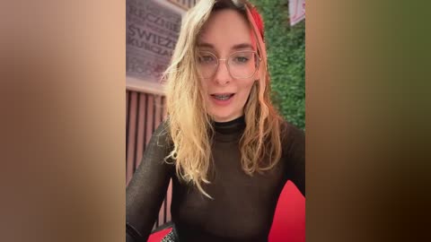 Media: Video of a blonde woman with wavy hair, wearing glasses and a black turtleneck, smiling, standing in front of a brick wall with greenery and a red sign in the background.