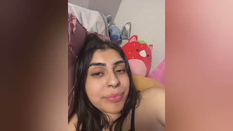 Media: Video of a young, light-skinned woman with long black hair, wearing a nose ring, lying on a bed with pink and purple pillows. A red plush toy and blue stuffed animal are in the background.