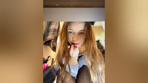 Media: A video of a young woman with long, light brown hair, wearing a denim jacket and a light blue shirt, licking her finger, taken from a smartphone. The background shows a messy room with clothes and a pink bag.