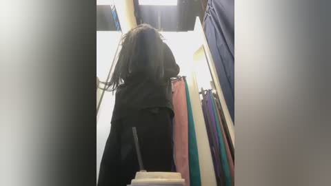 Media: Video of a person with dreadlocks, wearing a black jacket, standing in a narrow hallway with colorful clothing on hangers.