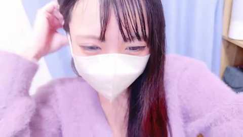 Media: Video of an East Asian woman with long, straight black hair, wearing a white medical mask, a lavender robe, and a concerned expression, indoors with blue curtains and a wooden shelf in the background.