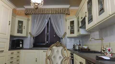 Media: Video of a classic kitchen with cream cabinets, a decorative chair, a beige countertop, a large window with sheer curtains, and a vintage chandelier.