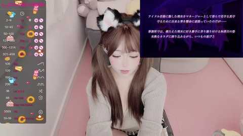 Media: Video of a young, fair-skinned girl with long brown hair, wearing cat ear headbands, a beige sweater, and sitting on a pink floor. Japanese text overlays the image.