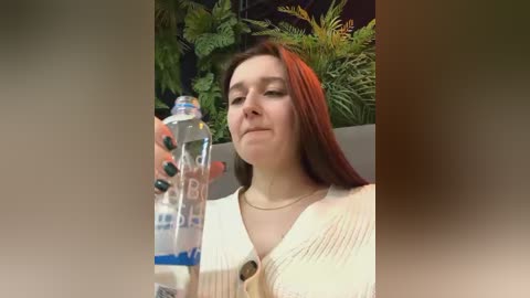 Media: Video of a fair-skinned woman with long red hair, wearing a beige sweater, holding a bottle of water in a modern indoor setting with green plants in the background.