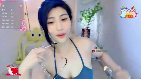 Media: Video of an East Asian woman with pale skin and dark blue hair in a ponytail, wearing a blue halter top, holding a phone. Background shows a green plant and a calendar.