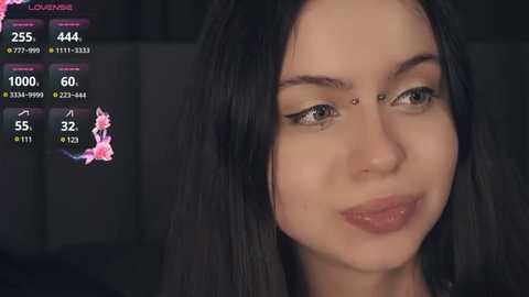 Media: Video of a young woman with long, straight black hair, fair skin, and green eyes, wearing subtle makeup and a nose ring. Background shows a dark, blurred room with a digital overlay displaying game statistics.