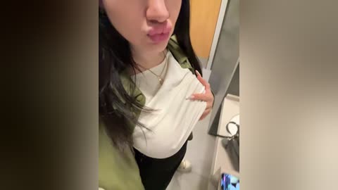 Media: A video of an East Asian woman with long black hair, wearing a white top and olive-green jacket, puckering her lips in a bathroom.