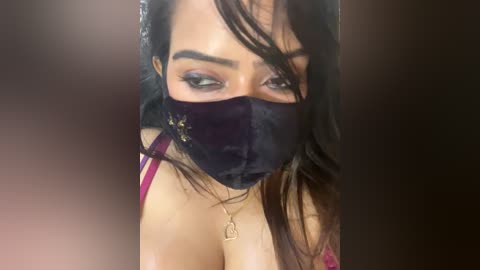 Media: Video of a woman with long dark hair, wearing a black face mask and pink bra, partially obscured by a dark background.