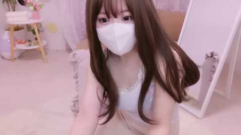 Media: Video of a young East Asian woman with long brown hair and a white face mask, wearing a white lace-trimmed camisole, kneeling on a soft carpet in a pastel-colored room with a white-framed mirror and a small wooden table.