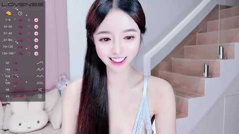 Media: A video of an East Asian woman with long black hair and fair skin, wearing a light blue lace top, smiling warmly in a modern, well-lit room with a staircase and plush teddy bear.