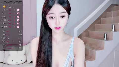 Video of a young, slender East Asian woman with long, straight black hair and fair skin, wearing a light blue lace bra, in a modern, softly lit room with beige walls and a wooden staircase.