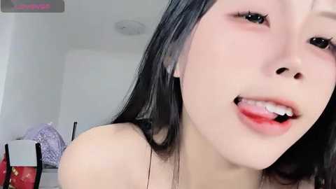 Media: Video of an Asian woman with long black hair, fair skin, and full lips, slightly open, looking seductively into the camera, in a modern, minimalist room with white walls and a purple couch.