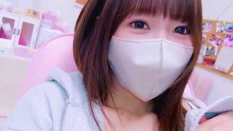 Media: Video of an Asian woman with long brown hair and blue eyes, wearing a white mask and light grey hoodie, sitting in a pink chair in a brightly lit room with white walls and shelves filled with toys and books.