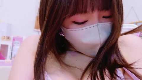 Media: A close-up video of a young Asian woman with long, straight brown hair wearing a white surgical mask, showing concern as she looks down. She has a soft, pale skin tone and is dressed in a light pink top. The background is blurred, with a hint of a pink and white setting.