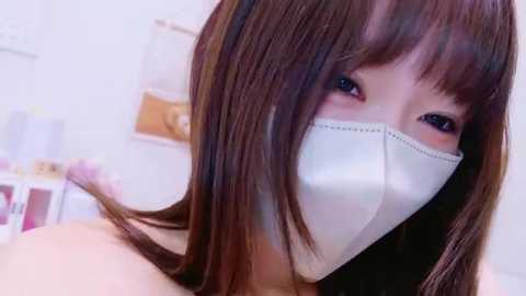 Media: Video of a young Asian woman with straight brown hair, wearing a white surgical mask, in a brightly lit room with a blurred background of shelves and boxes.