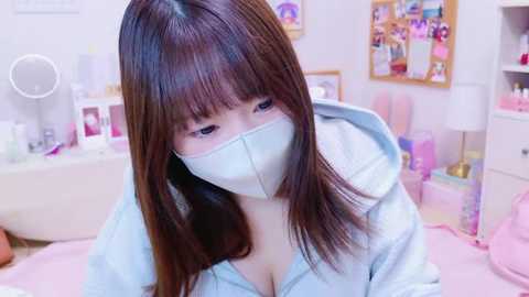 Media: A video of a young Asian woman with long, straight brown hair, wearing a light gray hoodie and a surgical mask, sitting in a pastel-colored bedroom. The room is decorated with pink and white accents and various personal items.