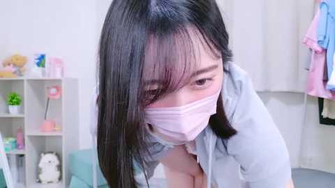 Media: A video of an Asian woman with long black hair, wearing a pink mask and light blue denim jacket, leaning forward in a brightly lit room with white walls, shelves, and a pink dress hanging.