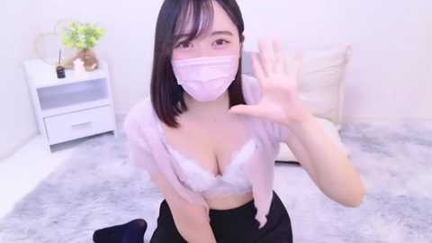 Media: Video of an East Asian woman with shoulder-length black hair, wearing a pink surgical mask and white lace bra, waving, in a modern, minimalistic bedroom with a white dresser and fluffy rug.