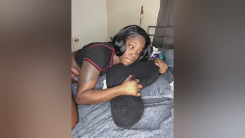 Media: Video of a black woman with medium-dark skin, shoulder-length black hair, lying on a bed, holding a black pillow, in a dimly lit bedroom.