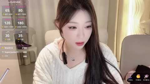 Media: A video of a young Asian woman with long black hair, fair skin, and a slender figure, wearing a white sweater, sitting in a modern room with a digital screen displaying streaming statistics.