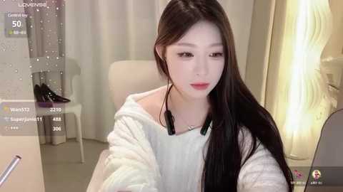 Media: Video of a young Asian woman with long black hair, wearing a white off-shoulder sweater, seated in a modern room with white walls and a lit lamp.